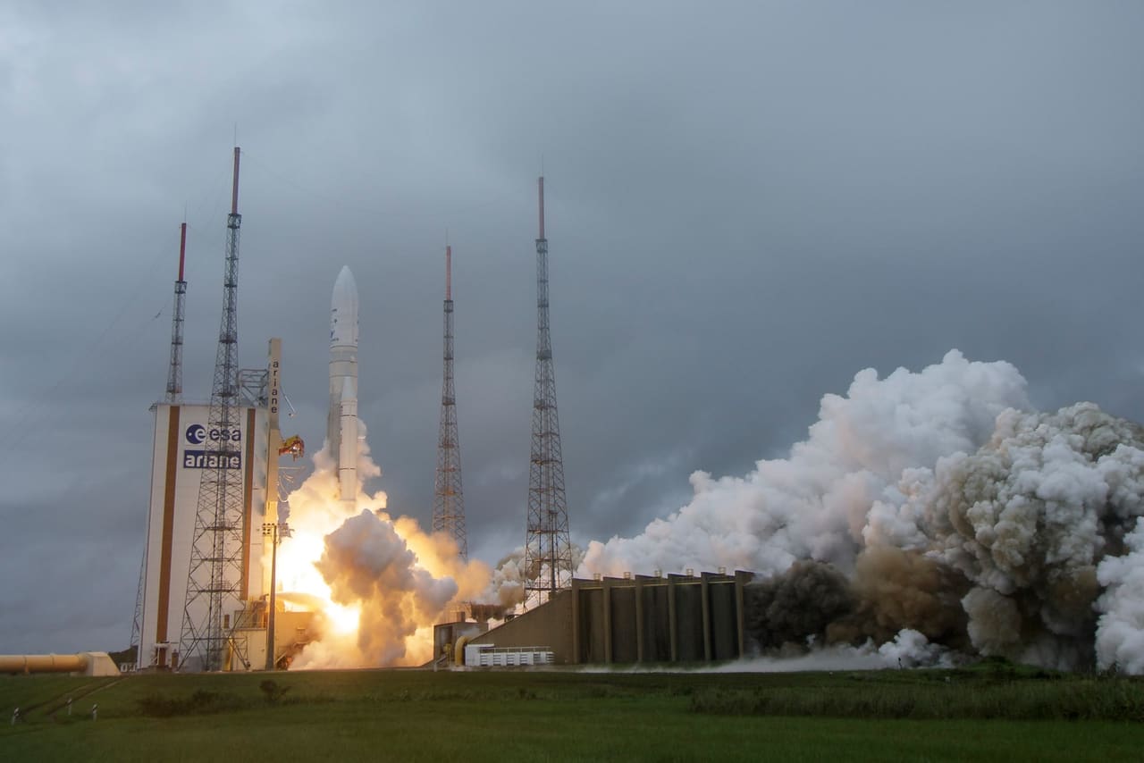 An Ariane 5 lifting off from Europe’s Spaceport in Kourou, launch tower and lightning masts on the left, the launch smoke cloud on the right