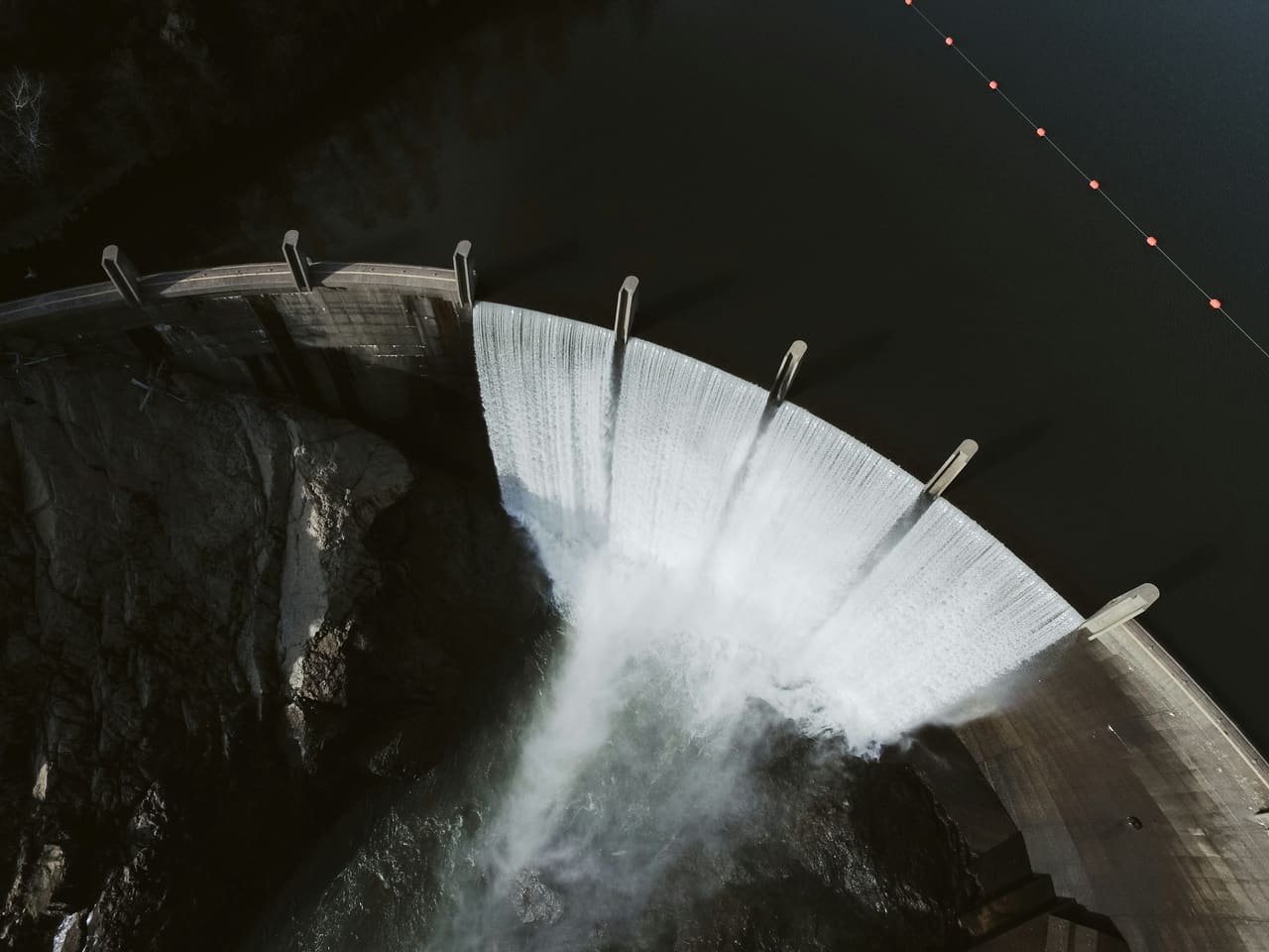 Senkrechte Luftaufnahme einer Staumauer, über deren Krone das Wasser hinwegstürzt.